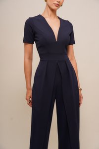 Marineblauwe jumpsuit van een gladde stof, met een diepe V-hals, korte mouwen en geplooide details aan de voorkant.