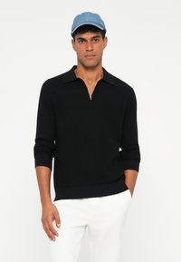 Sudadera negra con cremallera y cuello de polo, de punto texturizado y mangas largas, combinada con una gorra de denim azul claro.