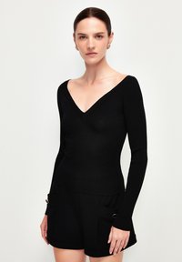 Schwarzes, geripptes Langarmtop mit V-Ausschnitt, körperbetontem Design und glatter Textur. Kombiniert mit schwarzen Shorts mit Taschendetails.