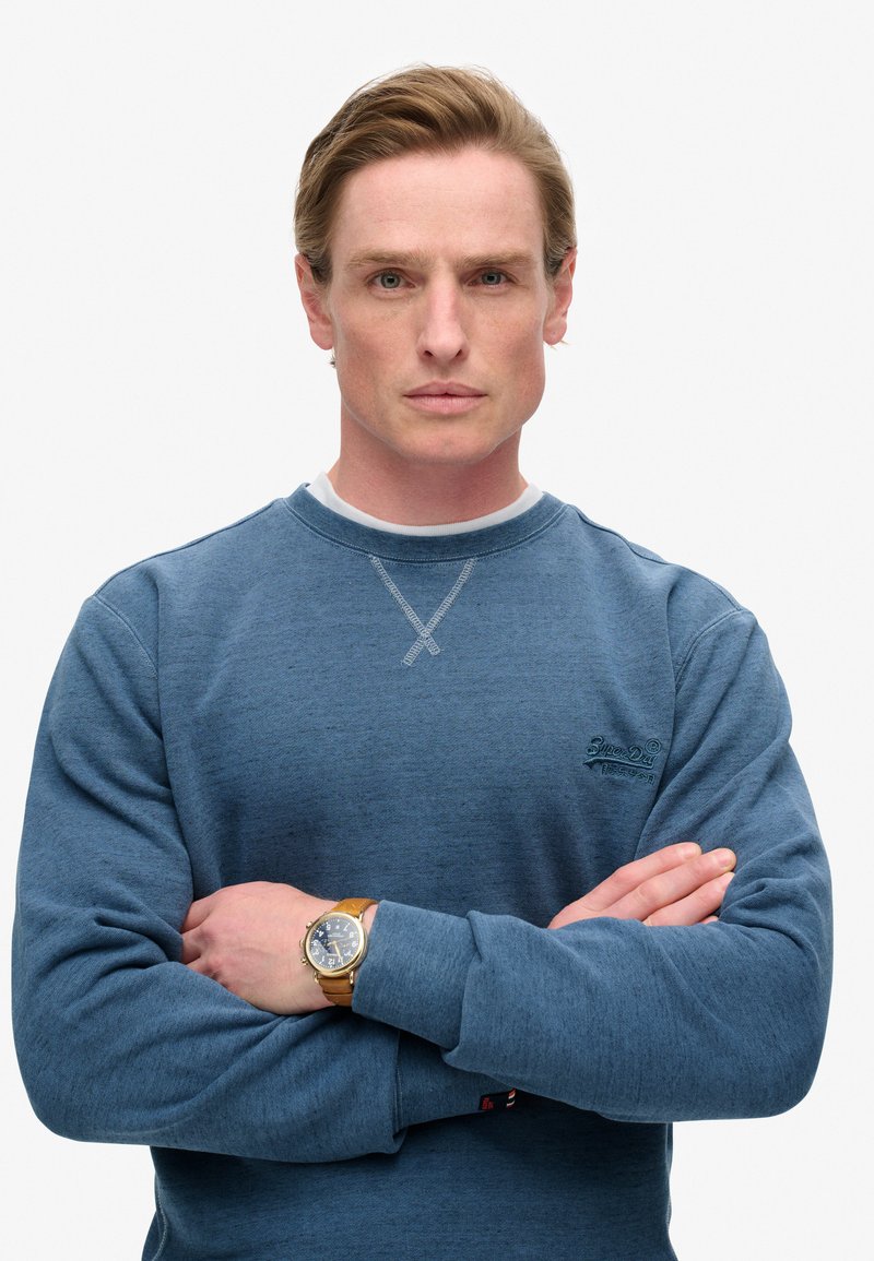 Hombre con pelo rubio que lleva un sudadera azul y un reloj en la muñeca, de pie con los brazos cruzados frente a un fondo blanco liso.