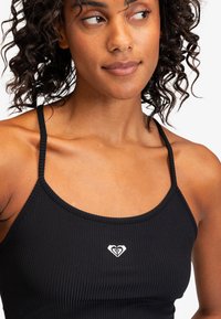 Svart ribbad crop top med tunna ännu band och en liten vit hjärtlogga i mitten. Tyget har en mjuk, texturerad yta.