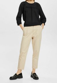 Schwarze strukturierte Bluse mit gerüschtem Ausschnitt, kombiniert mit beigen schmalen Hosen. Das Model trägt schwarze, polierte Slipper. Weißer Hintergrund.