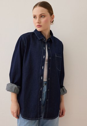 Jonge vrouw met het haar naar achter gebonden, draagt een donker denim overhemd met opgerolde mouwen over een wit topje en lichtblauwe spijkerbroek, staand tegen een witte muur.