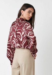 Woman with long dark hair wears maroon and white abstract patterned blouse and beige tailored pants, looking over her shoulder.