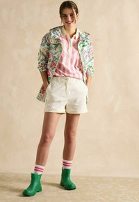 Jachetă cu model floral, polo cu dungi roz, pantaloni scurți albi, cizme de cauciuc verzi și șosete cu dungi. Culori vibrante și un look stratificat.