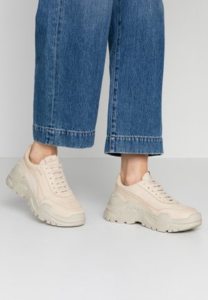 Person trägt cremeweiße klobige Sneakers und weit geschnittene, blaue, kurze Jeans vor einem einfarbigen hellen Hintergrund.