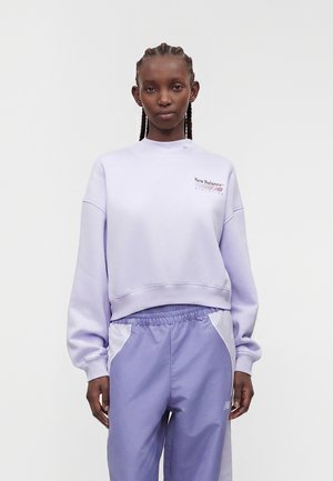 Ung person med flettet hår iført en lyse lilla New Balance sweatshirt og matchende sportbukser mod en ensfarvet baggrund.