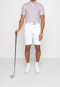 Shorts de golf blancs associés à un polo à manches courtes à motif de couleur violet clair. Comprend une ceinture argentée, des chaussures de golf blanches et un club de golf.