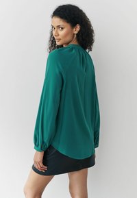 Bluză verde cu mâneci lungi, tiv curbat și detalii strânse la gât. Pur și simplu peste o fustă mini neagră. Textură a țesăturii netedă.