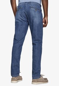 Hombre con pantalones vaqueros azules, de pie con la espalda mirando hacia adelante, zapatos casuales marrón claro y una camisa azul claro visible en la cintura.