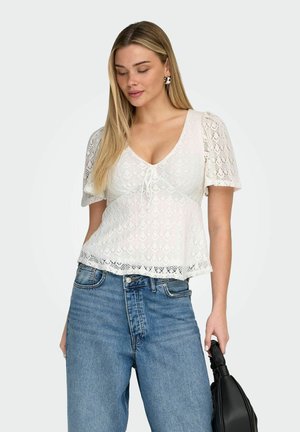 Kvinne med langt blondt hår iført hvit bluse med korte ermer i blonder og blå jeans, holder en svart håndveske ved siden av seg.