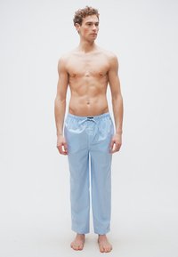 Νεαρός άνδρας όρθιος, ξυπόλητος, φοράει ανοιχτό μπλε και λευκή ριγέ πιτζάμα, κοιτάζοντας προς το πλάι με φόντο απλό.