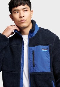 Fleecegefütterte Jacke in Dunkelblau mit blauen Einsätzen. Verfügt über eine Reißverschlusstasche auf der Brust, einen hohen Kragen und ein strukturiertes Material für Wärme.