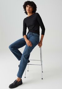 Zwart geribd longsleeve shirt gecombineerd met blauwe denim jeans. De jeans hebben een rechte snit en het model draagt zwarte schoenen terwijl ze zit.