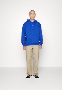 Sudadera azul con un bolsillo frontal y un logo blanco; combinada con pantalones beige y zapatillas negras con rayas amarillas. Fondo liso.