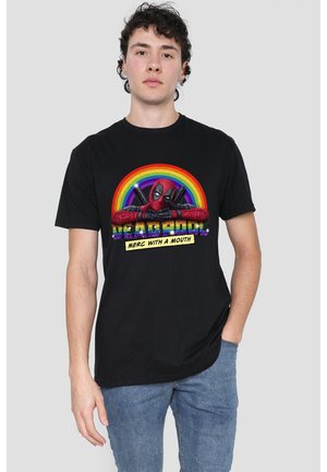 Junger Mann trägt schwarzes Deadpool-T-Shirt mit Regenbogen und Schriftzug „Merc with a Mouth“, kombiniert mit blauen Jeans, vor schlichtem Hintergrund stehend.