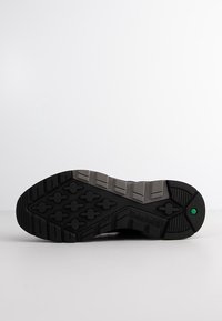 Semelle de chaussure Timberland noire et grise avec un motif de bande de roulement texturé et un petit détail hexagonal vert sur fond blanc.