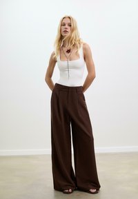 Femme blonde se tient à l'intérieur portant un haut sans manches blanc, un large pantalon marron, un collier et des sandales à bout ouvert devant un fond uni.