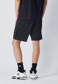 Pantaloni sportivi neri con bottoni laterali, lunghezza al ginocchio, realizzati in tessuto liscio. Indossati con scarpe da ginnastica bianche e calze nere a coste.