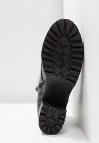 Semelle de botte noire avec des rainures profondes et des motifs floraux, taille 37, inclinée contre un mur et un sol blancs.