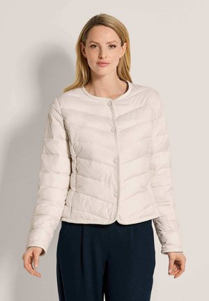 Frau mit glatten blonden Haaren, die eine hellbeige gesteppte Jacke und dunkle Hosen trägt, steht vor einem schlichten hellen Hintergrund.