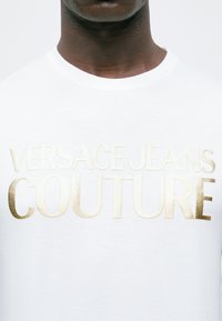 Tuvumā ir persona, kas valkā baltu krekli ar zeltīti izšūtu uzrakstu "VERSACE JEANS COUTURE" krūtīs.