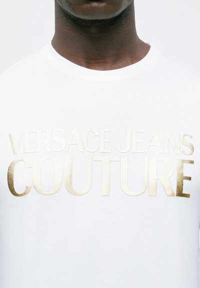 Крупен план на човек, носещ бяла риза с златно релефно лого "VERSACE JEANS COUTURE" на гърдите.
