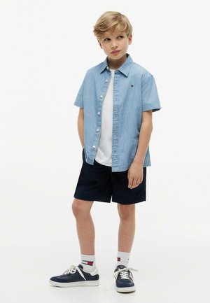Jongen met blond haar die een lichtblauw shirt met korte mouwen, een wit T-shirt, donkere korte broek, witte sokken en marineblauwe sneakers draagt, staand tegen een witte achtergrond.