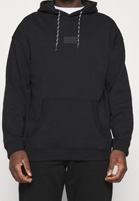 Black hooded sweatshirt made of cotton blend, features a front pocket, drawstrings with contrasting colors, and a textured logo patch.