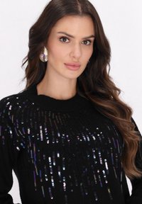 Schwarzer Pullover mit vertikalen Pailletten in verschiedenen Farben. Hat einen runden Ausschnitt und einen strukturierten Strickstoff. Auffällige Ohrringe mit lila Akzenten.