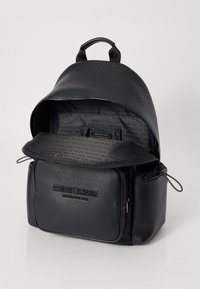Svart lær ryggsekk med teksturert ytre, glidelås på toppen, frontlomme og mønstret innside. Har "ARMANI EXCHANGE"-merke.