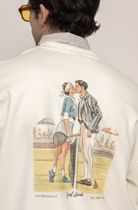Hvit sweatshirt med en fargerik illustrasjon av to tennisspillere som kysser, med racketer og et nett i bakgrunnen. Teksten "Lykke til" er inkludert.