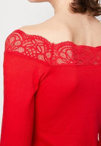 Röd off-the-shoulder topp med spetskrage, slät tyg och figursydd design. Spetsen har intrikata blommönster och scallopade kanter.