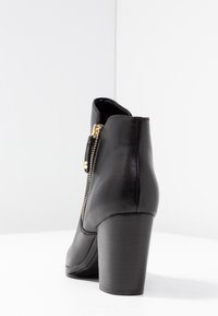 Botín negro de cuero hasta el tobillo con un alto tacón cuadrado, presenta un zipper lateral con herrajes dorados y una textura suave.