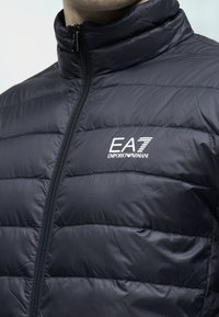 Jachetă căptușită neagră, cu guler înalt, tivit orizontal și fermoar frontal. Are logo-ul EA7 Emporio Armani pe piept.