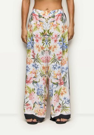 Mujer con pantalones blancos de pierna ancha con un patrón floral multicolor simétrico y borde negro en el dobladillo, de pie descalza.