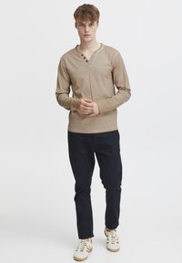 Beige pitkähihainen paita henley-aukolla, jossa on kolme pyöreää nappia. Yhdistetty tummiin housuihin ja valkoisiin tennareihin ruskeilla yksityiskohdilla.