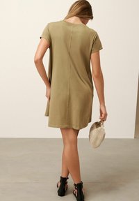 Robe courte olive à manches courtes avec une coupe ample, mettant en avant un détail de couture au dos. Associée à une pochette tissée beige et des sandales à lanières noires.
