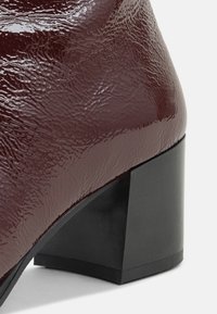 Bottine brillante bordeaux foncé avec surface texturée et talon bloc épais noir sur fond blanc.