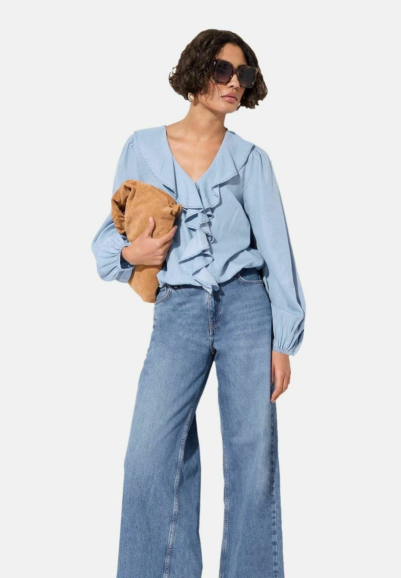 Blusa azul clara con escote con volantes, mangas largas de globo, combinada con jeans de pierna ancha. Sosteniendo un bolso de tela beige. Gafas de sol negras.