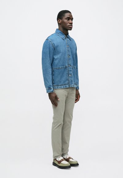 Mężczyzna stojący, ubrany w niebieską kurtkę jeansową, beżowe spodnie, białe skarpetki oraz zielono-brązowe mokasyny na jednolitym białym tle.