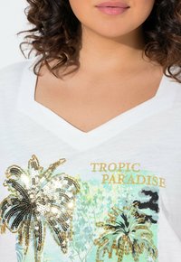 Bílý tričko s výstřihem do V, které má tropický design s flitrovými palmami a kovovým zlatým nápisem "TROPICKÝ RÁJ" na světle modrém pozadí.
