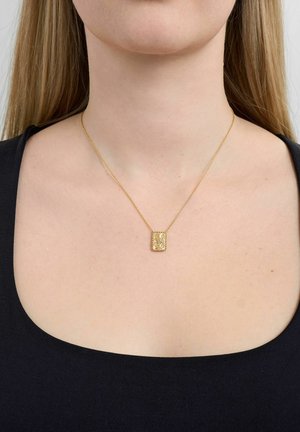 Kuldne ristkülikukujuline ripatsikaelakee peenete detailidega, kantud paljal kaelal musta sügava kaelusega topi kohal.