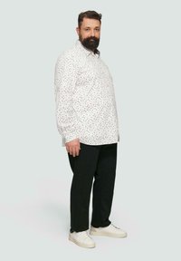 Weißes Hemd mit kleinen schwarzen und blauen Punkten, schwarze gestreifte Hose und weiße Turnschuhe. Das Model steht im Profil vor einem hellen Hintergrund.
