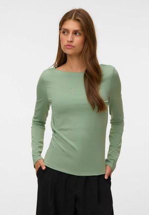 Femme aux longs cheveux bruns portant un top vert clair à manches longues et un pantalon noir, debout les mains dans les poches, regardant sur le côté.