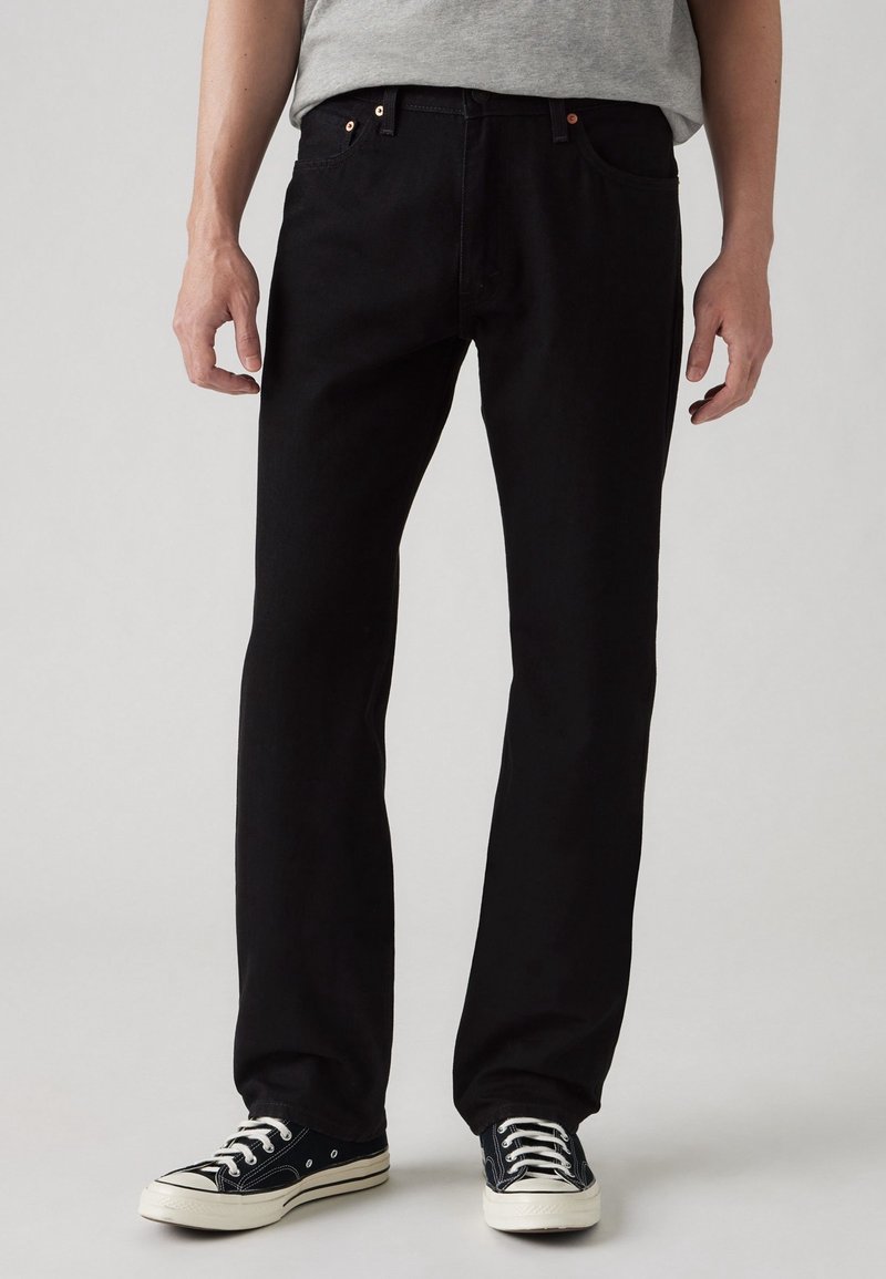 Černé džínové kalhoty s rovnými nohavicemi, jemným měděným kováním a hladkým povrchem, doplněné klasickými černými teniskami.