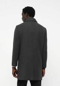 Cappotto in lana grigio a motivo houndstooth, con colletto, maniche lunghe e taglio dritto. Presenta un motivo testurizzato e dettagli metallici.