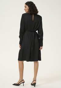 Robe noire avec manches longues, texture lisse et détail en forme de trou de serrure au dos. Comprend une ceinture à la taille et des accents de boutons sur les poignets.