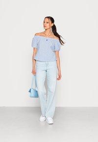 Blusa azul e branca de riscas, com mangas bufantes, sem ombros, combinada com jeans de perna larga azul claro e ténis brancos, segurando uma mala azul.