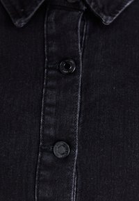 Μαύρο τζιν πουκάμισο με κουμπιά μπροστά, δίχρωμες ραφές και αιχμηρό γιακά. Τα κουμπιά είναι στρογγυλά και σκούρα, ταιριάζουν με το ύφασμα.